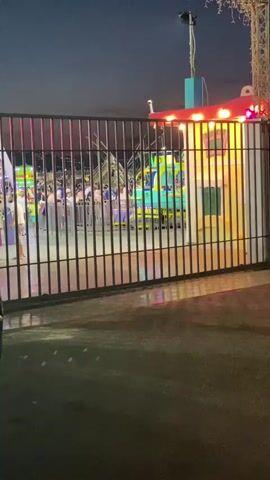 A black metal gate stands in the foreground, partially obscuring a scene at an amusement park. Through the gate, a brightly lit carnival ride is visible, possibly a kiddie coaster or a track ride with colorful cars. Several people are standing near the ride, some appearing to be waiting in line. The overall ambiance suggests an evening or nighttime setting, with artificial lights illuminating the attraction. The ground in front of the gate appears wet, reflecting the lights from the park.