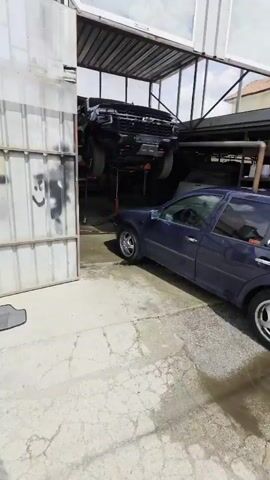 The video begins with a close-up of four car floor mats laid out on a concrete surface in front of a corrugated metal wall. The camera then pans to the right, revealing a dark grey sedan partially entering a garage with large metal doors. The garage door on the right is open, and a white door is visible behind it. Signs are posted above the garage opening. The sedan continues to move into the garage, and the camera follows, providing a side view of the car. The interior of the garage is dimly lit, with tools and equipment visible. A blue barrel with an orange cord coiled around it is positioned near the entrance. The car is now fully inside the garage, and the video ends.