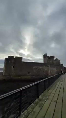 The video presents a series of shots from a coastal area, likely in Scotland given the presence of a Scottish flag atop a stone castle-like structure.  The initial view focuses on the fortress, which stands on a rocky shore. The overcast sky dominates the view with a diffused sun barely breaking through. The camera then moves to show the coastal landscape, featuring a calm, gray body of water meeting a dark, rocky shoreline. The next shot shows a wooden walkway with a vintage crane. The concluding shots shows a coastal scene with the gray water and overcast sky. The railings are made of metal and cable. The overall atmosphere of the video is somewhat bleak, reflecting the somber sky and muted colors of the landscape.
