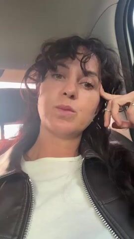 A close-up, selfie-style video shows a woman with dark, curly hair and fair skin. She is wearing a white t-shirt under a brown leather jacket with a silver zipper. Her hand, adorned with two rings, is resting on her chin and cheek, with her fingers near her mouth. She appears to be indoors, possibly in a vehicle, as indicated by the blurred background with what looks like a window and car interior elements. The lighting suggests it could be daytime, with natural light filtering in. The woman's expression shifts from a neutral gaze to a subtle smile and then a slight smirk, conveying a thoughtful or perhaps amused emotion. There is no visible text in the video. The provided location context of Bolshaya Rechka, Russia, places the scene in a specific geographic area.
