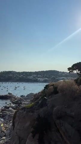 The video shows a body of water with many boats anchored. A rocky coastline is visible in the foreground, with vegetation on the rocks. In the background, a town or village is built on a hillside overlooking the water. The sky is clear and blue, with a faint ray of light visible. The camera pans slowly across the scene.