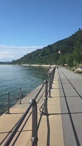 The video shows a scenic view of a lake on a sunny day. A paved walkway runs alongside the lake, separated by a railing. The walkway appears to be a promenade or a bike path, with some people visible in the distance, lounging or walking. The lake is calm and blue, with a gently sloping, tree-covered hill rising on the far side. The sky is clear blue with a few wispy clouds. The time of day appears to be late morning or early afternoon, given the bright sunlight and the long shadows cast by the railing. The location is Lochau, Austria, suggesting a picturesque European lakeside setting. The overall mood is peaceful and relaxing, characteristic of a beautiful summer day by the water. There is no visible text in the video.