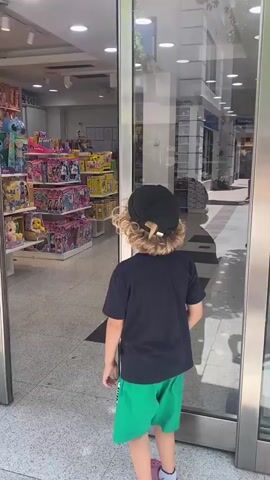 This video captures a young child, from behind, walking through a store. The child has blonde, curly hair and is wearing a black t-shirt, green shorts, and a black baseball cap. They are walking down a brightly lit aisle lined with shelves stocked with various items, appearing to be a toy store or a children's clothing store. The floor is tiled with a speckled pattern and has black rectangular mats placed strategically. In the background, more shelves and aisles are visible, extending deep into the store. A sign that reads "OVS" is visible further down the aisle, suggesting it's an OVS store. The lighting appears to be artificial, indicating the scene is indoors. The overall atmosphere seems casual and everyday, with no indication of a specific event or time of day beyond being indoors during business hours. The child's body language suggests movement and exploration.