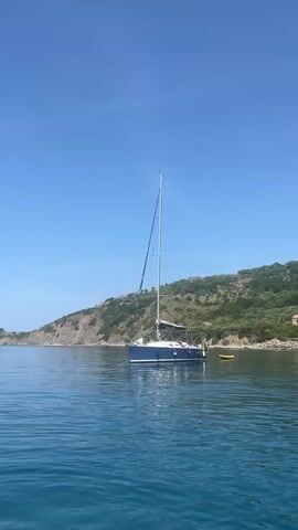 The video captures a serene coastal scene in Pollica, Italy, on a bright, sunny day. The sky is a clear, vibrant blue with only a small white cloud appearing briefly in the mid-section of the video. The water is remarkably calm, exhibiting shades of blue and green, suggesting clarity and depth.

The primary subjects are two sailboats anchored in the tranquil bay, positioned relatively close to the shore. The first boat to appear is blue, with its mast fully erected. A small, yellow buoy floats near its stern. As the camera pans from left to right, this blue boat eventually moves out of frame. The panning motion reveals more of the picturesque coastline.

The coastline consists of a lush, green hillside densely covered with trees and Mediterranean vegetation. Scattered across the hillside are several small buildings and houses, indicating some human presence. A distinctive small, rustic building with a red-tiled roof is visible on the lower slope of the hill. Further along the pan, a second sailboat, this one white, comes into view, also anchored with its mast up.

The scene conveys a sense of peaceful leisure, typical of a summer day by the sea. The calm water and clear weather suggest ideal conditions for boating or relaxing. There are no people visible, and no discernible activity other than the boats being anchored. No visible text is present in the video.