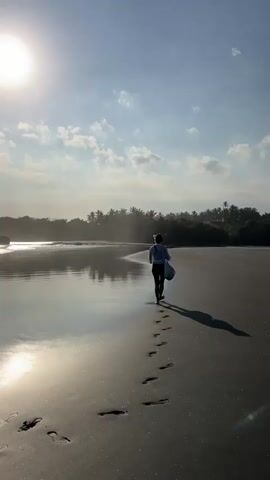 A barefoot woman with dark hair tied back, wearing a light-colored top, dark shorts, and carrying a bag over her left shoulder, walks away from the camera on a sandy beach. She follows a curve in the sand, leaving footprints behind her. To her left, the calm ocean reflects the bright sun and a clear blue sky with scattered clouds. Palm trees line the distant shoreline.