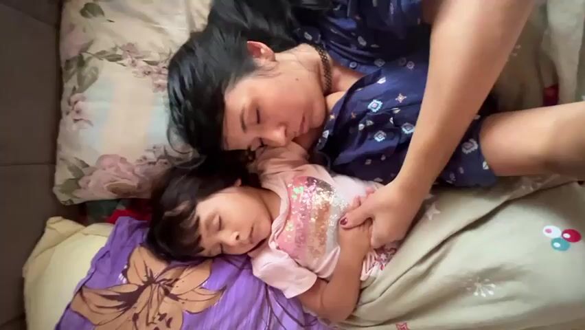 A top-down medium shot shows a woman and a young child sleeping in a bed. The woman, with dark hair, is lying on her side, facing the child. Her head is resting on a floral-patterned pillow, and she is wearing a dark blue patterned top. Her left arm is wrapped around the child. The child, who appears to be a girl with dark hair, is also lying on her side, facing the woman. She is wearing a pink t-shirt with a sequined pattern. Her head rests on a purple pillow with a floral design. The child's hands are clasped together, and the woman's hand is resting on top of them. They are covered by a beige blanket with a colorful circular pattern. The scene appears to be indoors, likely a bedroom, with soft lighting.