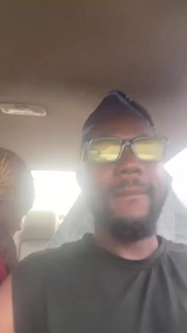 A selfie video is captured from inside a vehicle. The camera is focused on a man wearing a black tank top, a blue cap, and yellow-tinted glasses. He has a beard and is looking directly at the camera. His mouth is slightly open, and he appears to be speaking. In the background, a woman is visible, partially obscured, wearing a red top and a patterned headscarf. The interior of the car is dimly lit, with some light coming in from the windows. The video appears to be a casual recording, possibly during a car ride.