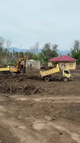 The video shows a construction site in Meunasah Lhok, Indonesia. The scene is dominated by a large pile of debris, primarily composed of logs and branches, with a thick layer of mud covering the ground. Two yellow construction vehicles are actively engaged in clearing the area.

In the foreground and middle ground, an excavator is seen digging into the debris pile. To its right, a yellow dump truck is positioned with its bed raised, receiving a load of mud and debris from the excavator. Further to the right, another yellow dump truck is parked.

In the background, there are several buildings. One prominent structure is a light yellow house with a red roof. Adjacent to it, there appears to be a smaller building under construction or in disrepair. Trees, some bare and others with foliage, are scattered across the landscape, and in the far distance, mountains are visible under an overcast sky. The overall impression is one of a cleanup or rebuilding effort following a significant event that has caused widespread debris and mud deposition. The time of day appears to be daytime, given the natural light. No people are clearly visible in the video. There is no discernible text in the video.