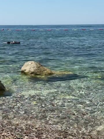 The video shows a clear, shallow body of water with a rocky seabed. A large, light-colored rock is partially submerged in the water, with its top portion visible. The water is calm and transparent, allowing the pebbles on the bottom to be seen. In the background, a few people are visible in the water, appearing to be swimming or engaging in water activities. A line of buoys marks an area further out in the sea. The sky above is a clear blue, suggesting a sunny day. The scene is likely a beach or a shallow coastal area.