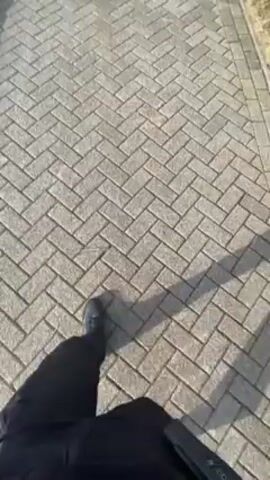 The video shows a person walking along a brick pathway. The pathway is paved in a herringbone pattern with gray bricks. The person is wearing black pants and black shoes. The shadow of the person and the camera is visible on the pathway. To the left of the pathway, there is a patch of snow. Beyond the snow, there is a partially snow-covered green space. To the right of the pathway, there is a row of tall, bare trees. The sky is blue with some light clouds. The camera moves forward as the person walks.