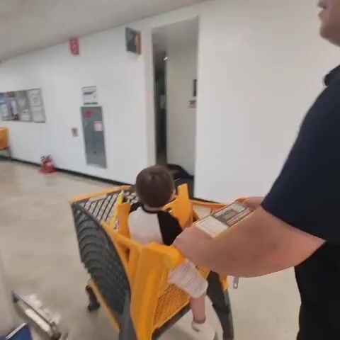 A first-person perspective video captures a scene in a brightly lit, indoor commercial setting resembling a supermarket aisle. In the foreground, a person's arm and hand are visible, pushing a yellow shopping cart with a child seated inside. The child, facing away from the camera, has dark hair and is wearing a black and white shirt and white shorts. The shopping cart has a separate compartment for a child. Further down the aisle, another yellow shopping cart is visible, with two other children and an adult. One child is interacting with items in their cart, and another child is walking behind them. The environment features white walls with framed posters and signage, a polished concrete floor, and fluorescent lighting. There are other shopping carts visible in the background, and a fire extinguisher mounted on the wall. The overall impression is of a family outing, likely grocery shopping.
