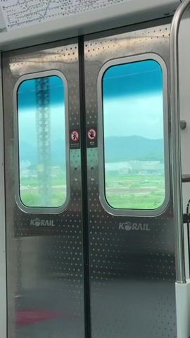 The video shows the interior of a train car. In the foreground are two stainless steel doors with windows. Through the windows, a blurred landscape of hills and fields can be seen. The Korail logo is visible on each door. A series of dots are patterned across the surface of the doors. There are two small signs with red circles and lines over icons of people on each door. The sky outside appears overcast.