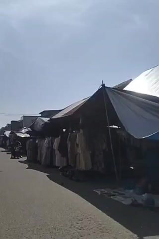 The video shows a street lined with market stalls on a sunny day. The stalls are covered with awnings made of corrugated metal and tarps. They are displaying clothing, primarily dresses and robes, hung on racks. A motorcycle with two people on it is seen riding down the street, passing by the stalls. In the background, there are buildings and power lines. The overall impression is of a bustling outdoor market.