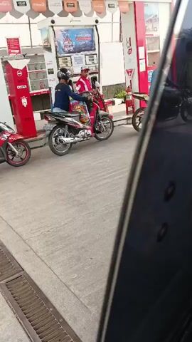 The video shows a gas station scene. Two people on motorcycles are at a gas pump, seemingly refueling. One person, wearing a white helmet and a dark shirt, is on a red scooter and appears to be operating the pump. Another person, wearing a dark helmet and a blue shirt, is on a red and silver motorcycle. A third person, wearing a red shirt and a red cap, stands between them, potentially a gas station attendant. The environment is paved and appears to be daytime. The camera is positioned from inside a vehicle, looking out.