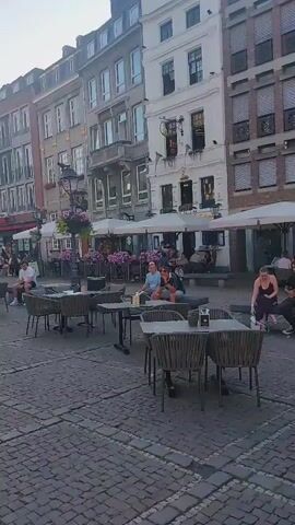 The video pans across a cobblestone square in front of a large, ornate building with multiple stories and many windows. In the foreground, there are several outdoor cafe tables and chairs. Some people are seated at the tables, dining and socializing. In the middle ground, a large, multi-tiered fountain is visible with a statue of a person on top. A man is sitting on the steps of the fountain, looking at his phone. Further in the background, more people are seen walking and sitting at tables. The overall atmosphere appears to be relaxed and pleasant, with a hint of historical architecture.