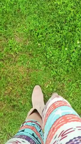 The video shows a first-person perspective of someone walking on a grassy area. The person is wearing tan fringed boots and colorful patterned pants. The ground is covered in green grass with some small white flowers and a few fallen leaves. There is a faint dirt path visible in the middle of the frame at one point. The camera angle is looking down at the person's legs and feet as they walk forward.