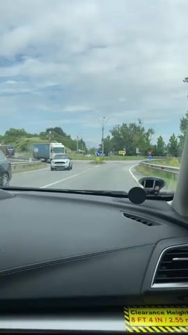 The video provides a first-person view from inside a car approaching and entering a roundabout in Drobeta-Turnu Severin, Romania. The car's black dashboard, windshield, and a phone mount are visible in the foreground. A yellow sticker on the lower right of the dashboard displays "Clearance Height 8 FT 4 IN / 2.5".

Outside, the scene is set on a cloudy day. As the car approaches the roundabout, a white Mini Cooper is seen to the left, followed by a white MAN truck entering the roundabout. The central island of the roundabout is covered in grass and small trees, featuring a blue circular sign with a downward-pointing arrow indicating the direction of travel, and yellow chevron signs. In the background to the left, a large cross-like structure is partially visible. The car then proceeds to enter and navigate the roundabout, turning right. Engine sounds and music can be heard in the background.