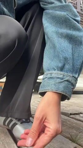 A person wearing a denim jacket and black pants walks towards two chairs on a wooden deck. A small dog sits on a cushion on one of the chairs. The person sits down in the other chair, and the dog jumps onto the person's lap. The person pets the dog. The environment is an outdoor deck with a building in the background.