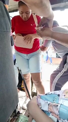 The video shows the interior of what appears to be a public transport vehicle in motion. A woman wearing a red long-sleeved shirt and light blue denim shorts is standing near the center of the frame. She is holding a small, light-colored purse and appears to be taking something out of it. Several other people are crowded around her; a person in a gray shirt stands to her left, and another individual wearing a green shirt is partially visible to her right. The person recording the video is holding several bills of money. Through the open doorway of the vehicle, a street scene is visible with a mix of people and storefronts.
