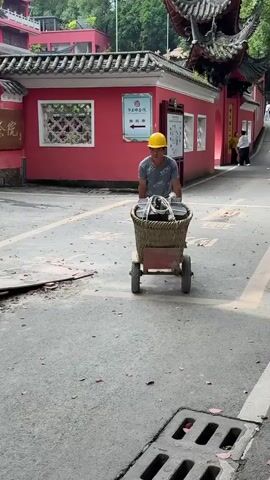 A man is seen pulling a wheeled cart that is carrying a large woven basket filled with stacks of what appear to be black tiles or building materials. The man is wearing a yellow hard hat, a light blue t-shirt, black pants with yellow stripes, and gloves. He is walking along a paved road beside a building with a red exterior and traditional Chinese architectural features. Construction materials and fencing are visible in the background, indicating ongoing work. The road has a storm drain and is scattered with red flower petals. The man is focused on pulling the cart.