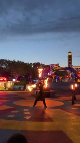 A group of performers are engaged in a fire-dancing show in an outdoor plaza as dusk settles. The main performer in the center, dressed in dark clothing, wields two flaming batons, creating arcs of fire. Behind them, a large bonfire burns, spewing sparks. In the background, large, colorful, abstract sculptures, possibly part of a decorative installation, are visible, illuminated by colorful lights. A second performer, also in dark attire, can be seen to the right, also holding a flaming object. The ground of the plaza features a patterned design with red and yellow elements. The sky is a deep blue, indicating the transition from day to night. Buildings and trees are visible in the background.