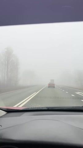 The video shows a view from inside a car driving on a highway in very foggy conditions. Another car is visible a short distance ahead, and a large truck appears further in the distance, its headlights visible through the fog. Bare trees line the side of the road. The road surface appears wet. The windshield is visible in the lower portion of the frame, and water droplets are present on its surface. The car's dashboard is also visible. The overall scene indicates limited visibility due to the heavy fog.