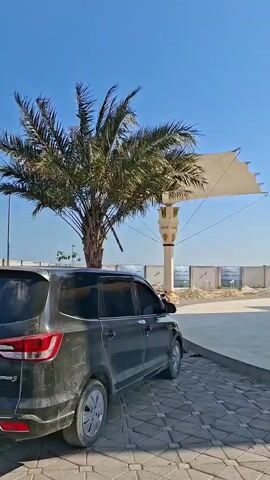The video opens with a view of a car parked on a paved area. In the background, a structure resembling the Kaaba is visible, featuring black walls with gold trim. A large white canopy supported by pillars shades the area. The environment appears to be outdoors under a clear blue sky with some clouds. The camera pans, showing more of the Kaaba replica, decorative pillars, and a fence in the distance. Palm trees are also present in the scene. The paving stones are arranged in a patterned design. The video concludes with a wider shot of the area under the canopy.