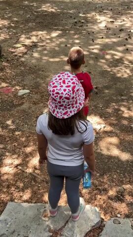 A person records two children from behind in a forest setting with adventure park elements. The child on the left wears a white t-shirt and a red and white patterned bucket hat. The child on the right wears a red t-shirt and green shorts. They stand on a raised concrete surface, facing away from the camera, looking into the forest. In the background, tall trees are visible, with wooden platforms, rope bridges, and ladders built into them, part of an adventure course. The ground is dirt with scattered pine cones and shaded areas.
