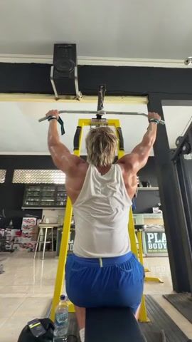 A person with blond hair is performing a lat pulldown exercise in a gym. They are seated on a black bench, facing away from the camera, and gripping a silver bar attached to a yellow weight machine. The person is wearing a white tank top and blue shorts. Their back muscles are clearly defined and appear to be sweaty. In the background, there are shelves with various items, a black speaker mounted on the ceiling, and a sign that says "PLAY BODY WORK HARD." A water bottle and a black bag are on the floor next to the bench. The lighting in the gym is bright, and the floor is tiled.