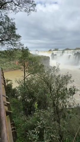 A wide shot captures a vast, multi-tiered waterfall under a cloudy sky. The water is a murky brown, churning as it plunges over a cliff face. Lush green trees and foliage frame the foreground, with some branches obscuring parts of the waterfall. A brown railing is visible on the left side of the frame. The scene appears to be a natural landscape, likely a national park or protected area.