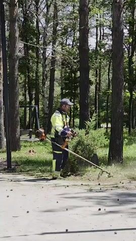 A man in a yellow and blue uniform with reflective stripes operates a string trimmer in a wooded area. He is wearing a baseball cap and protective eyewear. The trimmer has an orange engine and a long shaft. The ground around him is covered with pinecones and scattered debris. The background consists of tall trees with sunlight filtering through their leaves, indicating it is daytime. The overall impression is of yard maintenance or groundskeeping in a park or natural setting. There is a swing set visible in the far left background. The weather appears to be clear and sunny.