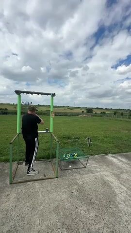 The media file shows a man participating in a shooting sport at an outdoor range. The man, dressed in a black t-shirt, black pants with white stripes, and a baseball cap, stands in a green metal shooting stand. He appears to be holding and aiming a firearm. Beside the stand, a green metal rack holds several clay targets.

The setting is a grassy field under a partly cloudy sky. The clouds are white and fluffy, suggesting a daytime scene, possibly late morning or afternoon. The sky is a vibrant blue. In the background, there are hints of agricultural fields and possibly some distant structures.

The overall atmosphere seems focused and active, consistent with a sporting event. The man's posture suggests concentration as he prepares to shoot. There are no visible signs of distress or overt emotion, indicating a calm and controlled environment.

The location context provided is "Фельдмаршальский, Russia." This information, combined with the visual cues of the open field and the type of shooting range, suggests a rural or semi-rural setting in Russia.

There is no discernible text visible in the media file.
