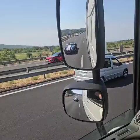 The video shows a view from inside a vehicle, likely a truck, driving on a highway. The primary focus is on a blue and white classic sports car, possibly a Shelby Cobra, traveling in the same direction. The sports car is visible in the side mirror and also in the windshield's reflection, indicating it's directly in front of or alongside the camera vehicle. The highway is multi-lane with other vehicles present, including cars and a larger truck in the distance. The scenery outside the vehicle is a sunny day with green hills and trees lining the road. The interior of the truck is visible, with a dashboard, mirrors, and a GPS device.