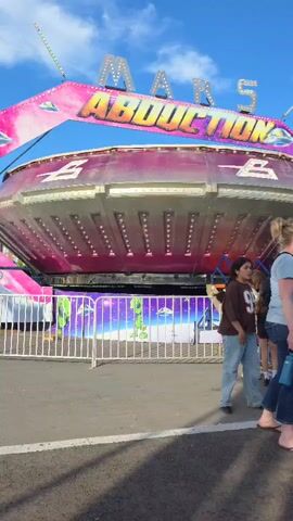 The video shows the entrance to a carnival ride called "Mars Abduction." The ride is a large, round structure with lights and an awning that says "MARS ABDUCTION." In front of the ride, there is a fence with images of aliens and UFOs. Several people are standing in line to enter the ride, and others are walking past. The sky is blue with some clouds, and the ground is paved.