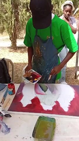 A person wearing a green shirt, black cap, and paint-splattered apron is painting on a canvas. The canvas is mostly red with a large white shape in the center that appears to be the beginning of a flower. The painter holds a palette of paints in their left hand and uses a brush in their right hand to apply paint to the canvas. On the table next to the canvas are several paint containers, brushes, and a cloth. Behind the painter, another person wearing a straw hat and jeans stands watching. The scene takes place outdoors under a tree, with other people and objects visible in the background.