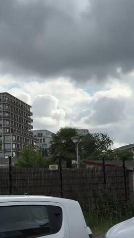 A vertical video captures an outdoor scene with a cloudy sky overhead. In the background, a tall, modern building with many windows stands on the left. To the right of it are other buildings, some appearing to be apartment complexes, and a palm tree. A fence constructed of bamboo-like material runs across the mid-ground of the frame, with a sign attached to it. A white car is partially visible in the foreground at the bottom of the frame. A bird is seen flying in the sky, captured by the camera at multiple points throughout the short clip. The overall environment appears to be urban or suburban.