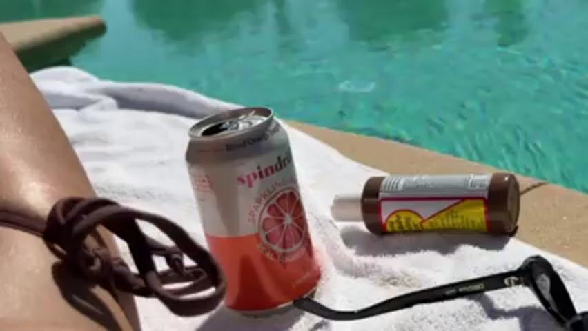 A sunbathing scene shows a partially visible person wearing a brown swimsuit lying next to a pool. A white towel sits on the pool’s edge, holding an open can of Spindrift sparkling water (Blood Orange flavor), a bottle of sunscreen, and a pair of sunglasses. The pool water is turquoise. The scene is brightly lit.
