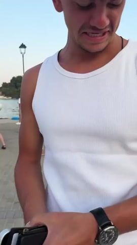 The video begins with a close-up of a young man's face. He is wearing a white tank top and has short, dark, messy hair. He appears to be speaking or reacting to something off-camera. The scene then shifts to a wider shot of a waterfront area. Several people, dressed in white, are gathered on a paved walkway next to a large boat named "MAKEDONIA PALACE." Some people are standing, others are talking in small groups. The boat has multiple levels and appears to be preparing for or hosting an event, with lights and screens visible. In the background, there is water and some greenery on what appears to be a hillside. The final shot is a low-angle view of someone's legs and feet, wearing white sneakers and a white skirt, walking on a paved surface. The overall impression is a gathering or event by the water, possibly a party or tour, with people dressed in white.