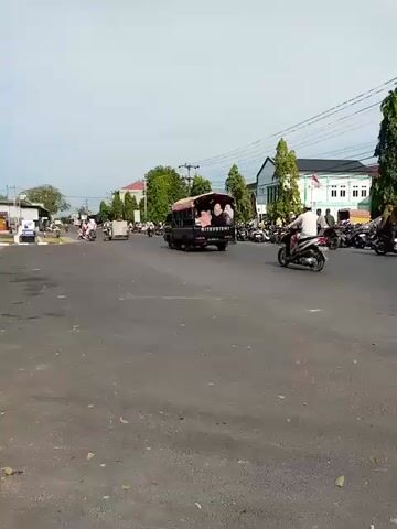 The video shows a street scene with a few vehicles and people. There is a dark-colored van in the middle of the road, and a motorcycle rider in front of it. On the right side of the road, there are many parked motorcycles and a few people standing around. In the background, there are some buildings and trees. The sky is clear and the sun is shining. The video is shot from a low angle, giving the impression of being on the street level. The time stamp indicates that the video was recorded on January 5, 2024, between 00:00 and 00:05.
