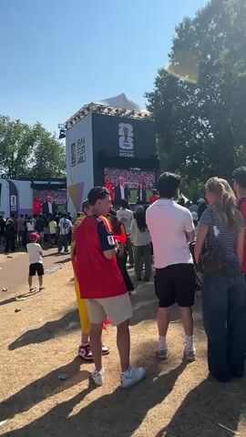A group of young people are gathered outdoors on a sunny day, watching a large screen displaying the FIFA World Cup 2022 logo. Some are wearing sports jerseys, and one person in the foreground is holding a red vuvuzela. The screen shows a man in a suit speaking. The environment appears to be a park or festival ground with dry grass and trees in the background. The people are casually dressed and appear to be enjoying the event.