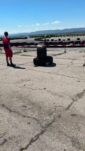 This is a vertical, outdoor, daytime video clip of a person in red shorts and a red shirt standing on a cracked asphalt surface, looking towards a race track. Two tires are stacked in the foreground, with two water bottles on top of the bottom tire. The track is lined with black tire barriers, some of which have red and white tape on them. Several cars are visible on the track in the background, along with a few people wearing high-visibility vests. The sky is clear and blue, with mountains visible in the distance.