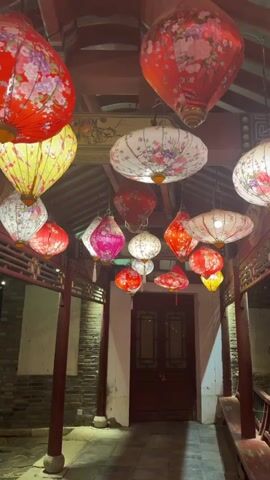 The video displays an outdoor walkway decorated with hanging floral lanterns. The walkway is covered by a traditional Chinese-style roof, supported by red wooden pillars on each side. The floor of the walkway is paved with stone tiles. At the end of the walkway is a dark wooden door with a locking mechanism. Lanterns of various shapes, colors, and floral patterns hang from the roof, illuminating the walkway. The surrounding walls appear to be made of gray brick. The scene suggests a festive or cultural atmosphere.