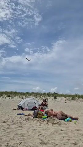 The video shows a beach scene with several people and objects. In the foreground, a person is lying on the sand, wearing a patterned swimsuit. To their left, a white tent is set up, with a person sitting in a chair next to it. There are several beach towels and bags on the sand. In the mid-ground, a person is sitting in a red folding chair, wearing a black hoodie and shorts, looking at a smartphone. Another person is visible in the distance, lying on the sand. Further back, there are several tents and beach umbrellas, as well as other people in the distance. The sky is partly cloudy, and there are seagulls flying overhead. The overall impression is a busy beach on a cloudy day.