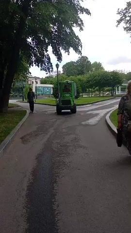 In a park setting, a person is walking along a paved path. To their right, a small green vehicle, possibly a utility vehicle or a mini-tractor, is being operated by someone. The vehicle is moving in the same direction as the person. The park features lush green trees and grass, with a paved walkway and a path for vehicles. In the background, there are buildings and more trees, suggesting a suburban or urban park. The lighting indicates it is daytime, with the sky appearing cloudy. The overall atmosphere seems calm and ordinary, likely a typical day in the park. There is no visible text in the frame. The person walking appears to be wearing casual clothing, and the vehicle operator is partially visible. The scene captures a moment of everyday activity in an outdoor public space.