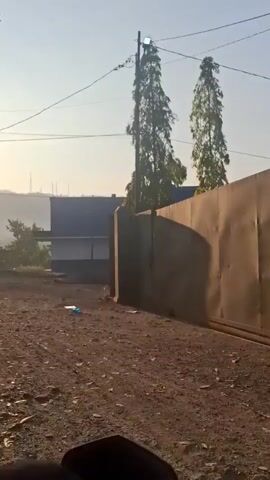 The video begins showing a dirt road with some debris. A building with a fence is visible to the right. Two tall trees stand behind the fence. The sun is rising, casting a warm glow and creating lens flares. The camera then pans to reveal a mosque with a dome, followed by a small building surrounded by greenery. The video appears to be filmed from a moving vehicle, possibly a motorcycle, as a mirror and handlebars are partially visible in the foreground. The overall environment suggests a rural or suburban setting in the early morning.