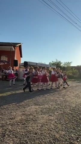 A group of children in traditional folk costumes are dancing outdoors in front of a building. The children, a mix of boys and girls, are arranged in rows and appear to be performing a choreographed dance. They are wearing brightly colored outfits, with the girls in red and white dresses and the boys in shirts and pants, some with vests. The background includes a building with a red facade, a white tent or stage structure, and trees. The ground is covered in gravel and dirt, and shadows are cast on the ground, indicating it is late afternoon or early evening. The sky is clear and blue, with a faint haze of dust visible in the air around the dancers, suggesting movement and activity. Some adults are visible in the background, near the building. The overall scene depicts a cultural performance or celebration.