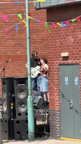 A man and a woman are standing on a platform with musical equipment. The man is holding a white electric guitar and appears to be playing it. The woman is standing close to him, looking towards him and appears to be singing into a microphone. They are both dressed casually. Behind them is a brick wall decorated with colorful triangular flags. To the left of the platform are large speaker cabinets. To the right is a gray door. The overall scene suggests an outdoor musical performance or rehearsal.