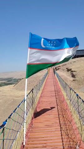 A video shows a flag of Uzbekistan blowing in the wind on a suspension bridge. The bridge has a wooden walkway and multi-colored netting for railings. The background is a dry, hilly landscape with a clear blue sky.