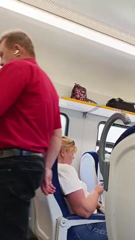 The video shows the interior of a train. In the foreground, a woman with blonde hair tied up with an orange scrunchie is seated, looking down at something in her hands. She is wearing a white t-shirt and blue jeans. Her left arm is resting on the back of her seat. In the background, another passenger, whose back is to the camera, is leaning their head against the window and looking out. This passenger has curly, graying hair and is wearing a tan shirt. Outside the window, green trees and a cloudy sky are visible, suggesting the train is moving through a rural area. Overhead lighting fixtures are visible on the ceiling of the train carriage.