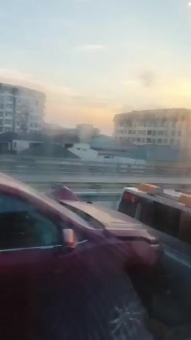 The video shows a view from inside a vehicle moving along a highway. The sky is a pale blue with hints of yellow and orange from the setting sun. Buildings, some appearing to be apartment complexes and smaller houses, are visible in the distance. Highway barriers line the road, and other vehicles, including cars and a white van, are seen driving in the opposite direction. The foreground is dominated by the interior of the vehicle from which the video is being recorded, with a reflection of the outside world visible on the window. The overall impression is that of a journey through a somewhat urban or suburban landscape at dusk.